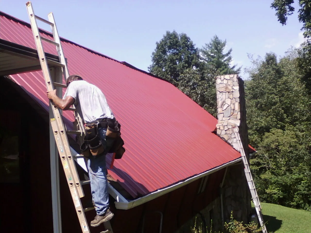 Roofer on a ladder beside a red metal roof and stone chimney — Soffit & Fascia Repair by Roofing Brothers in Eastport